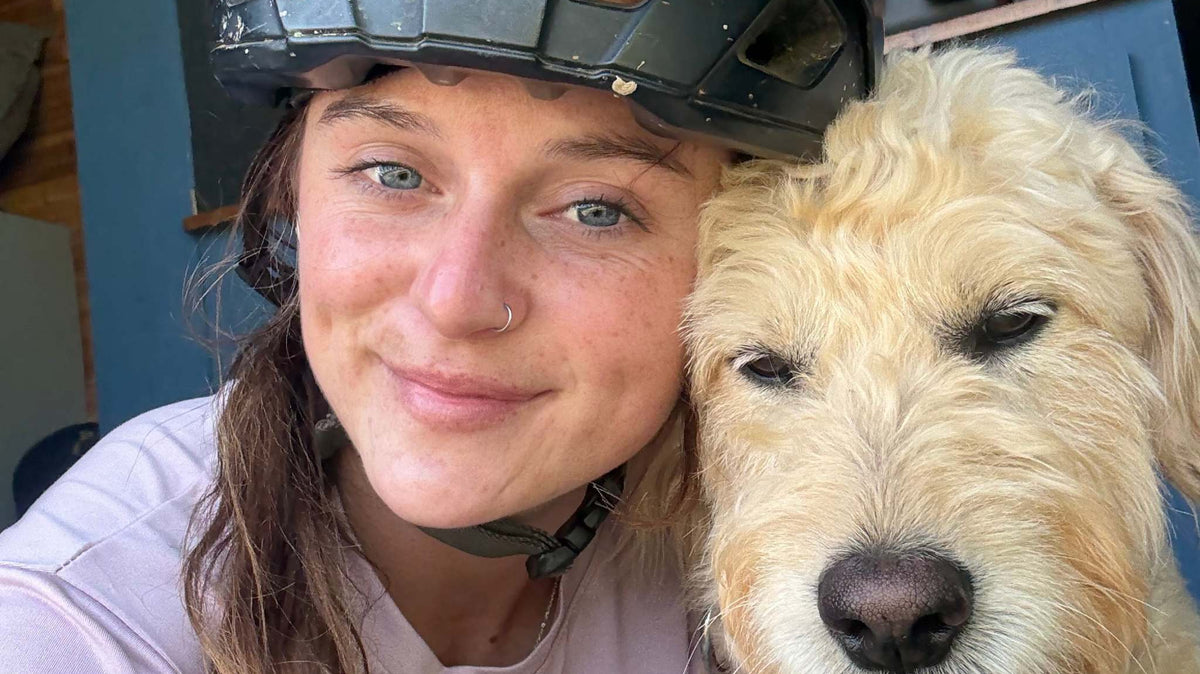 Woman wearing a bike helmet and Trail Dog Collab mountain bike jersey posing with a light-colored dog