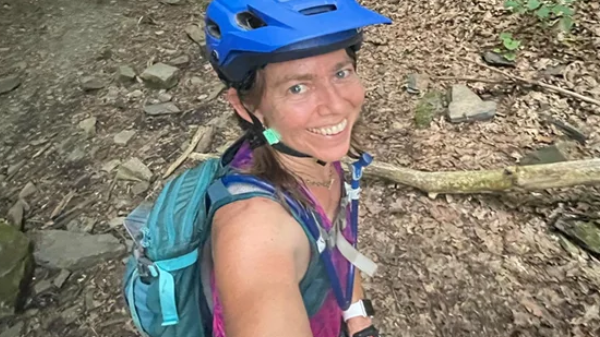 Woman mountain biker wearing blue helmet and backpack smiling, taking a selfie in a forest trail near a tunnel during the Wilderness 101 Marathon.