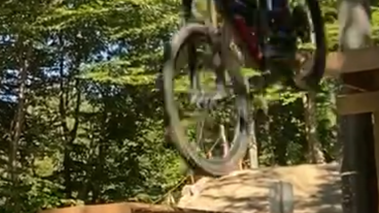Mountain biker performing a jump on a dirt trail in a forested downhill bike park, emphasizing deliberate practice and skill improvement.