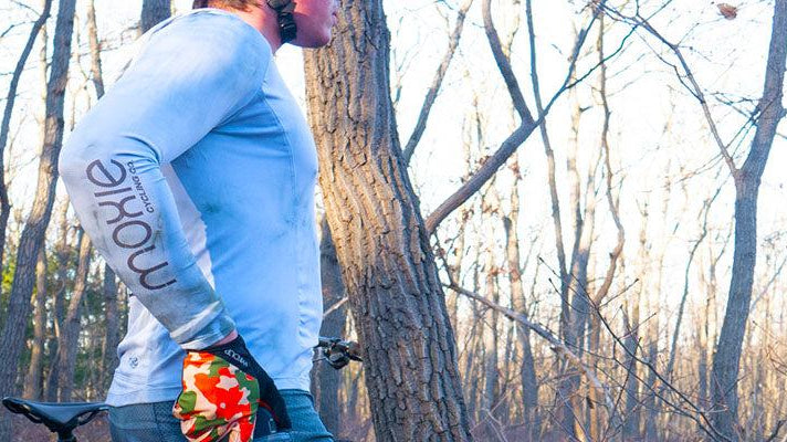 Person wearing Moxie Cycling long sleeve jersey riding a bike in a wooded trail, promoting inclusive unisex cycling apparel from the Cycling For All collection.