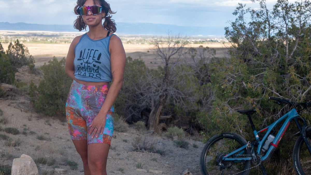 Women wearing colorful gravel shorts with bike in desert landscape.
