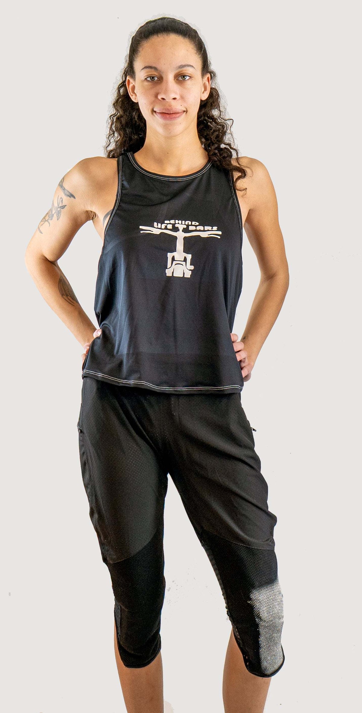 Woman wearing Moxie Tech Tank breathable mesh cycling jersey with "Life Behind Bars" graphic on mountain bike trail