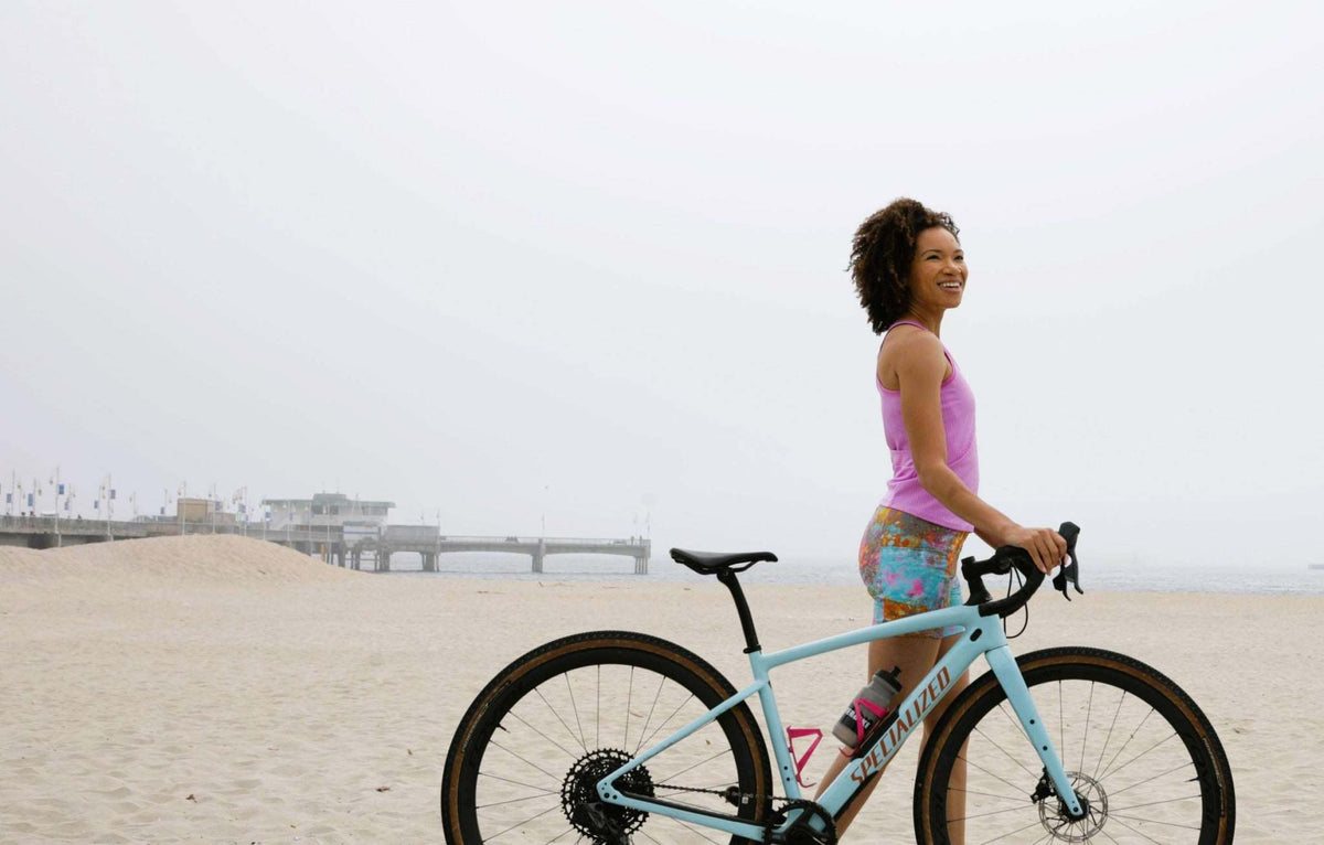 Woman wearing colorful gravel shorts standing with a bicycle on the beach.