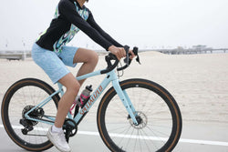 Woman riding a light blue Specialized gravel bike on a beachside path wearing a long sleeve cycling jersey and blue cycling shorts