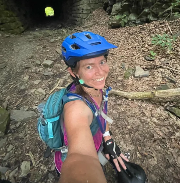 Woman mountain biker wearing blue helmet and backpack smiling, taking a selfie in a forest trail near a tunnel during the Wilderness 101 Marathon.