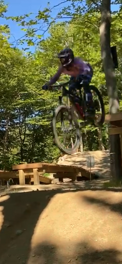Mountain biker performing a jump on a dirt trail in a forested downhill bike park, emphasizing deliberate practice and skill improvement.