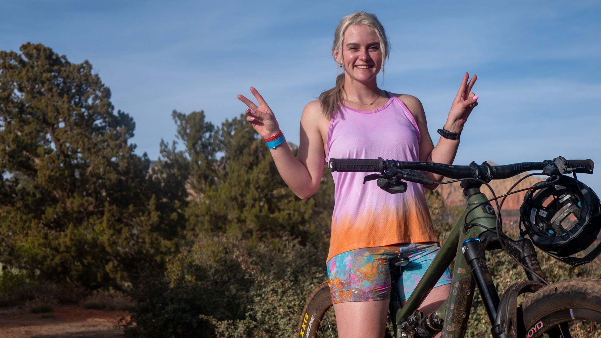 Smiling female mountain biker in colorful outfit making peace signs with bike in Sedona spring landscape