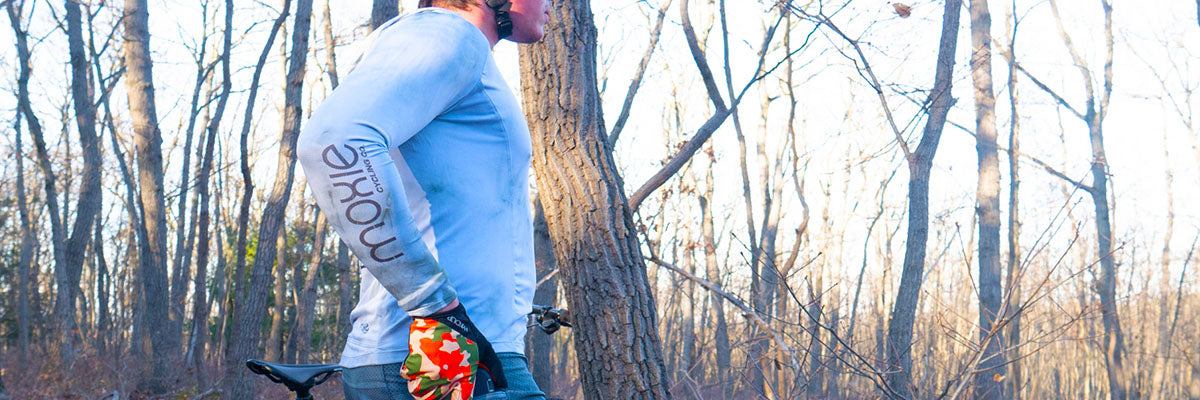 Person wearing Moxie Cycling long sleeve jersey riding a bike in a wooded trail, promoting inclusive unisex cycling apparel from the Cycling For All collection.