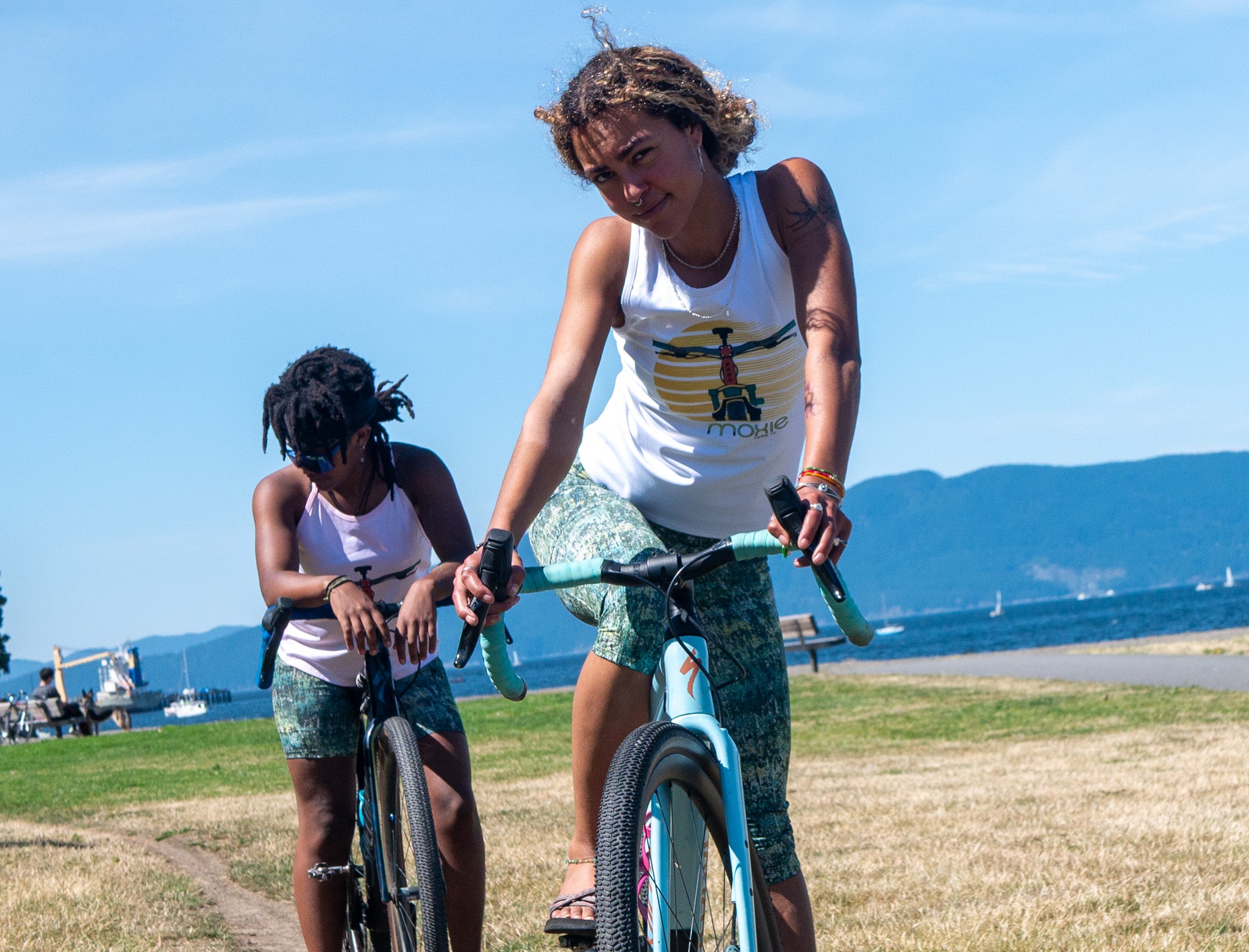 Two women riding bicycles outdoors on a grassy path near the water, with mountains and blue sky in the background. One woman is wearing a Moxie sleeveless jersey with a graphic print and Moxie knickers, while the other wears a white tank top and matching green patterned shorts.