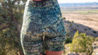 Women's gravel bike shorts with sewn-in chamois, green pattern, and elastic waistband in scenic outdoor setting.