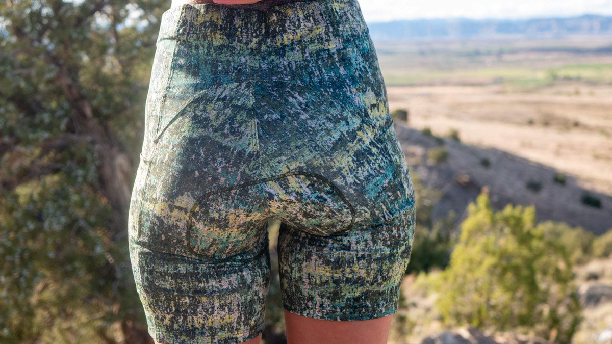Women's gravel bike shorts with sewn-in chamois, green pattern, and elastic waistband in scenic outdoor setting.