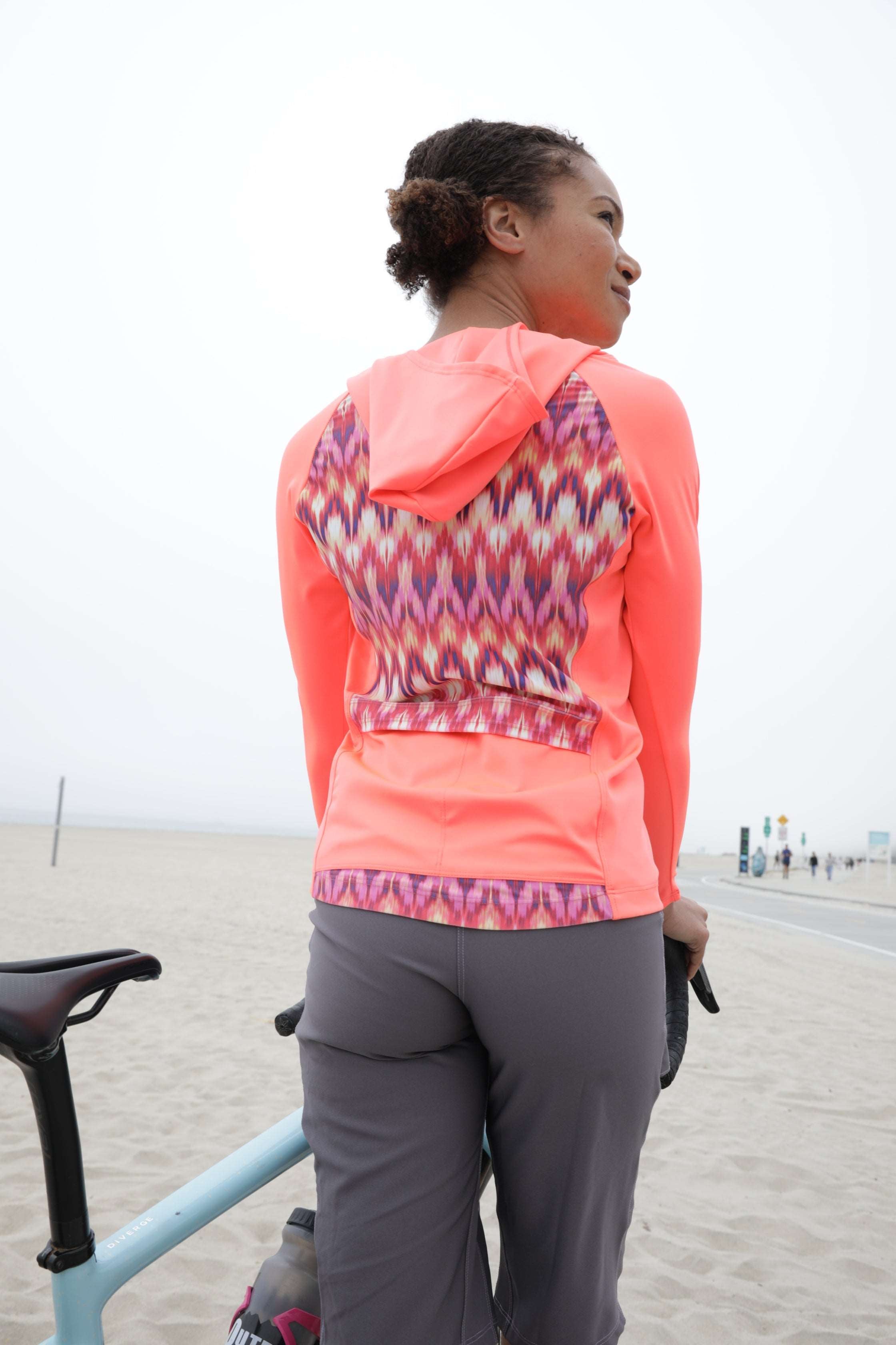 Moxie Tee Heavier Weight hoodie in vibrant design worn by a person standing with a bicycle on a beach background.