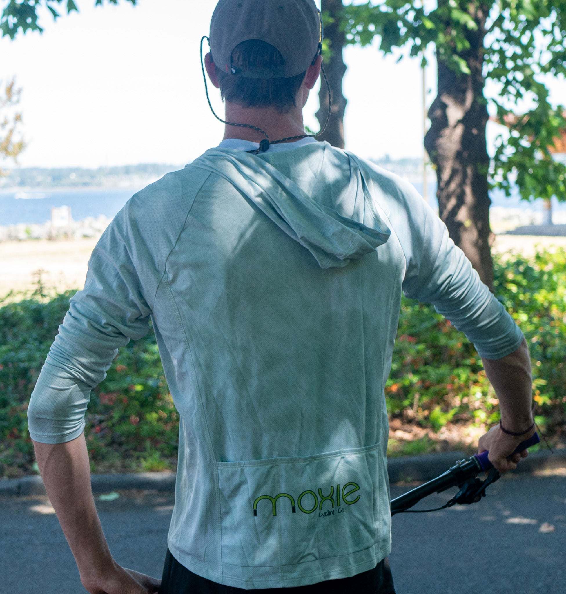 A man viewed from behind wearing a sage green hooded mountain bike jersey with 2 back pockets