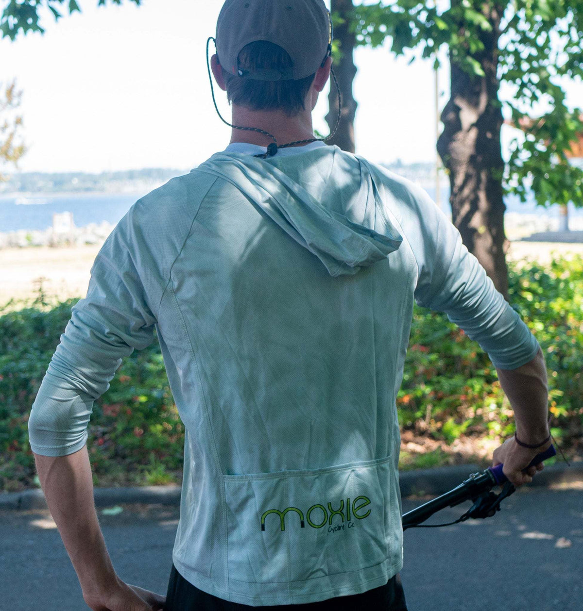 A man viewed from behind wearing a sage green hooded mountain bike jersey with 2 back pockets
