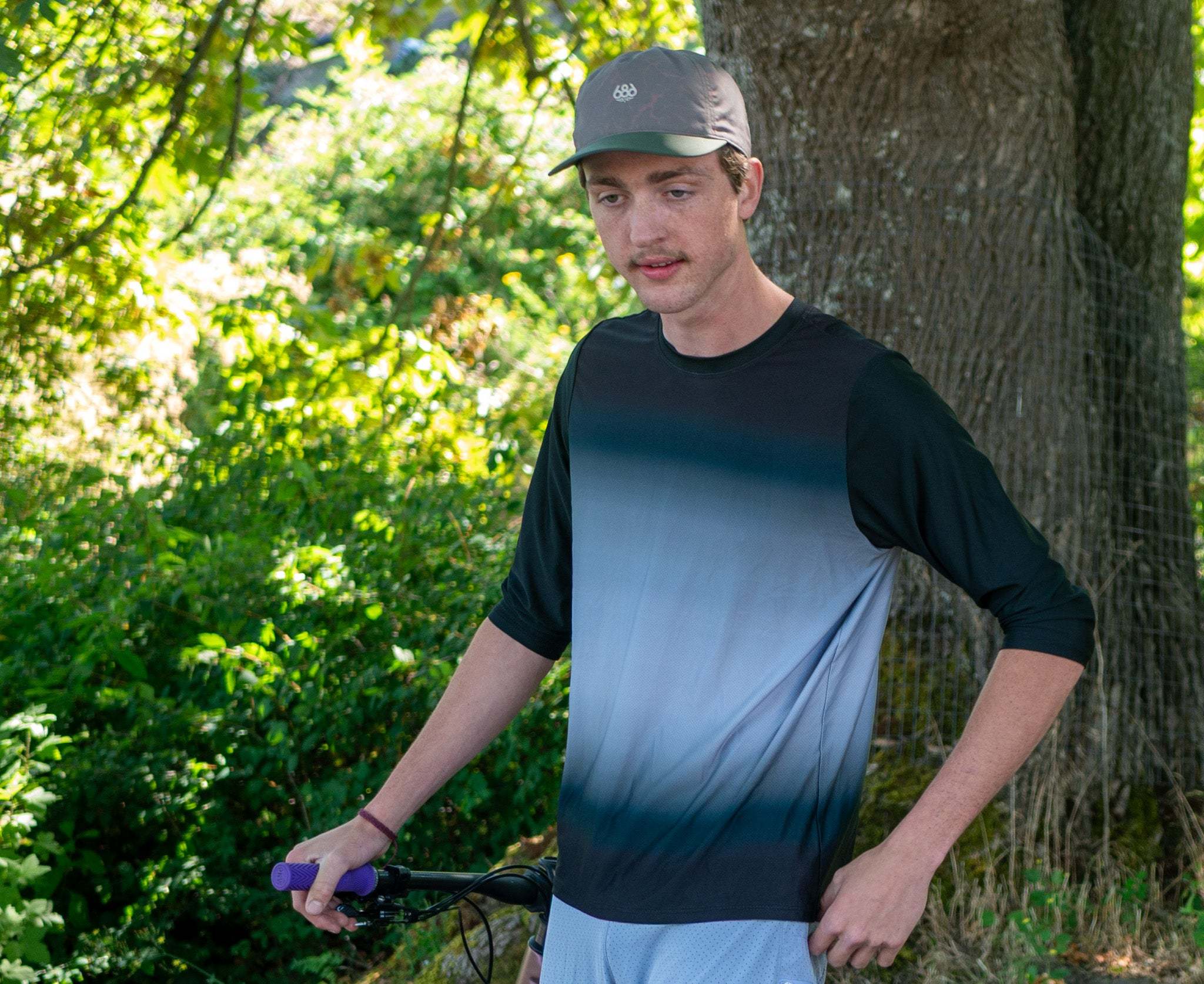 A man wearing a dark gray and light blue gradient 3/4t-sleeve mountain bike jersey in gray fade by his mountain bike