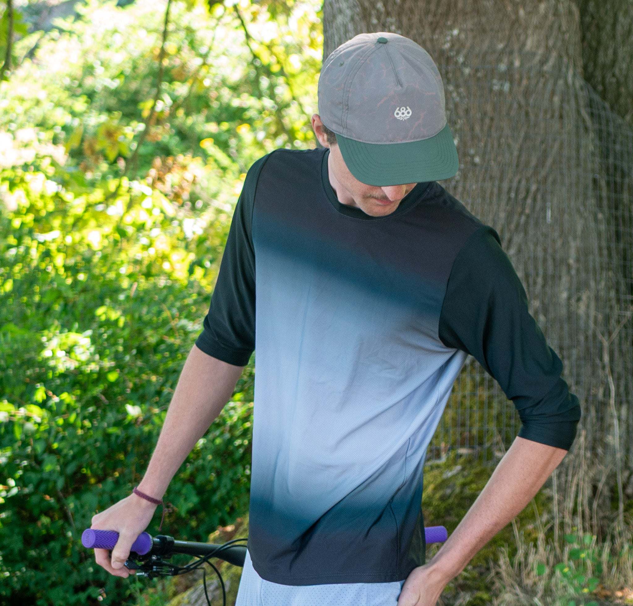 A man wearing a 3/4 sleeve mountain bike jersey by Moxie Cyclingt with a black-to-light-blue gradient body and solid black sleeves, paired with a gray cap, standing outdoors near a tree and holding a bicycle handlebar.