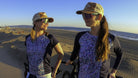 Women wearing Moxie Tee Jersey Blues, cycling on a beach at sunset.