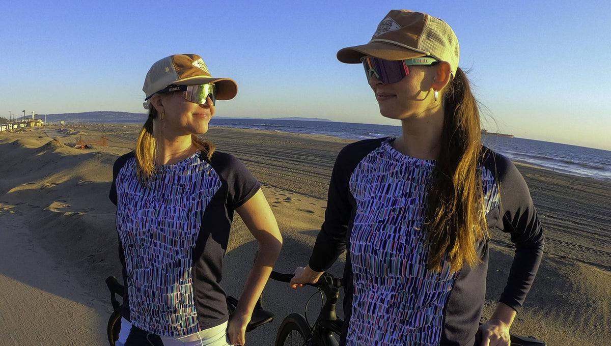 Women wearing Moxie Tee Jersey Blues, cycling on a beach at sunset.