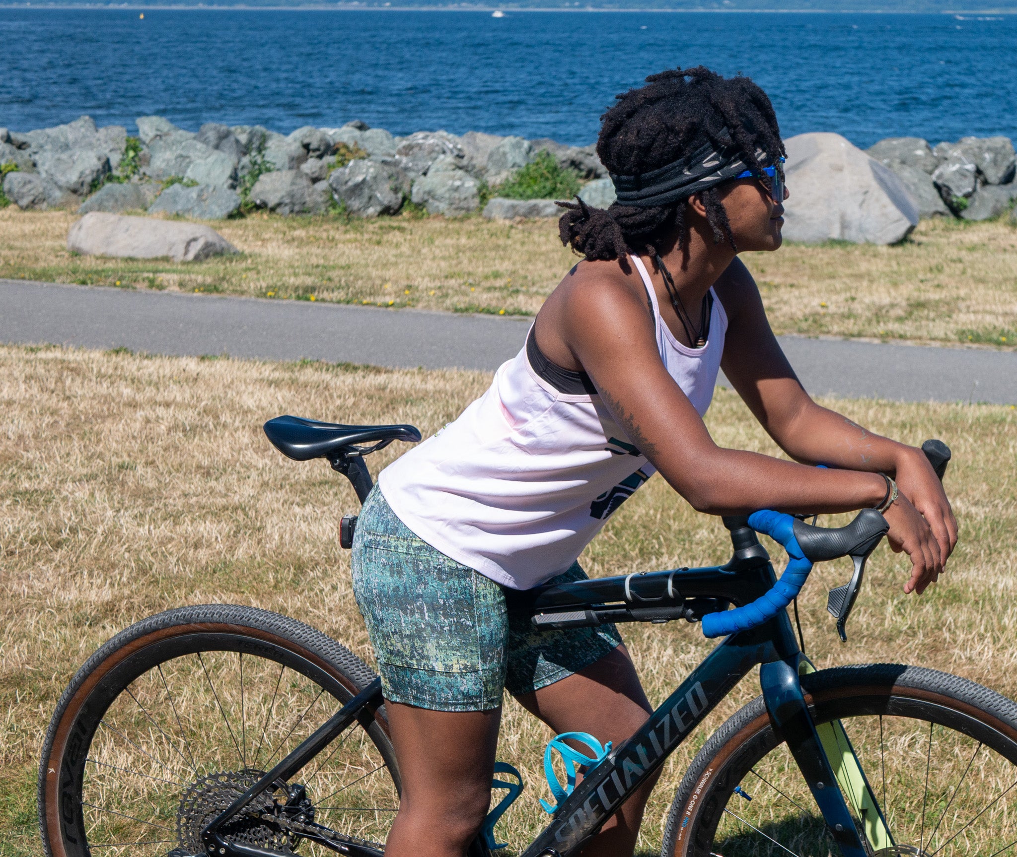 A woman riding a black Cannondale road bicycle outdoors on a grassy path near the ocean, wearing a Moxie cycling women's jersey, patterned shorts, and a black headband.