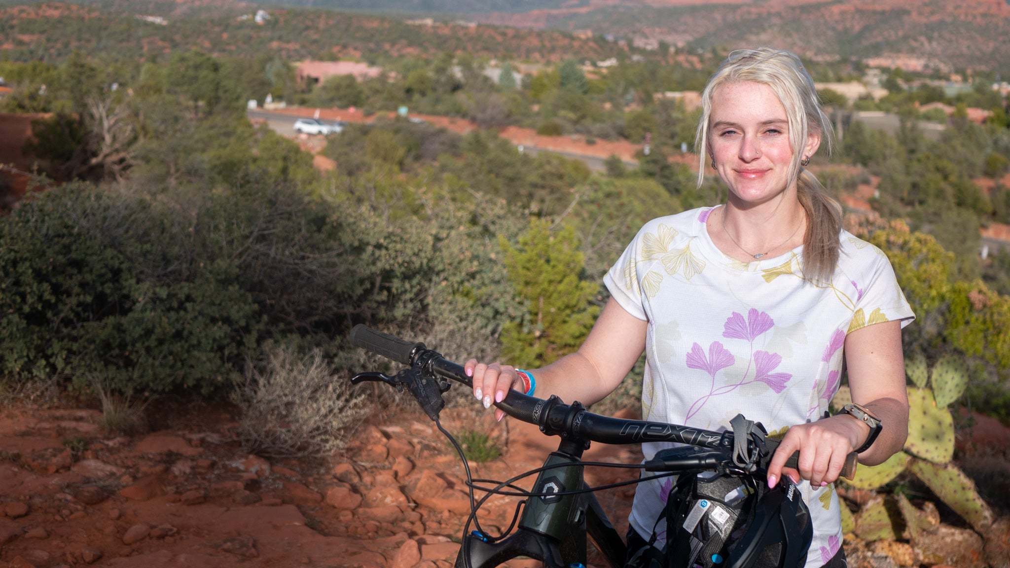 Woman wearing Moxie Tee Jersey Moss Floral shirt holding a mountain bike outdoors in a desert landscape