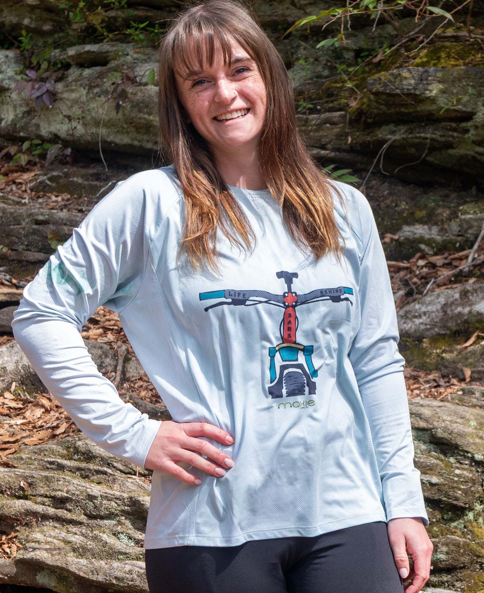 Person wearing a light green mountain bike hoodie with a graphic design, standing outdoors among rocks and foliage.