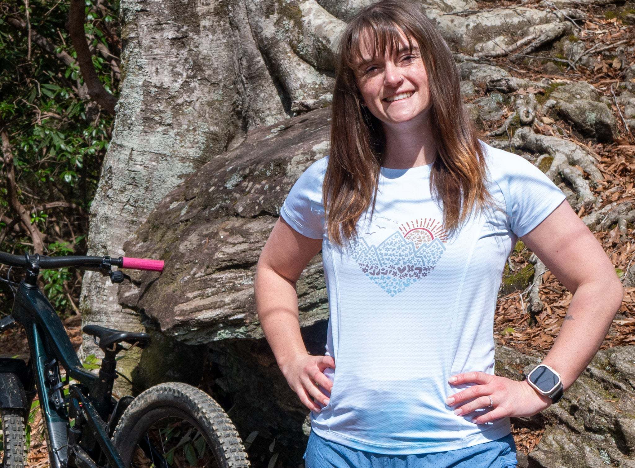 Person wearing a light blue t-shirt with a design, standing outdoors near a tree and bicycle.