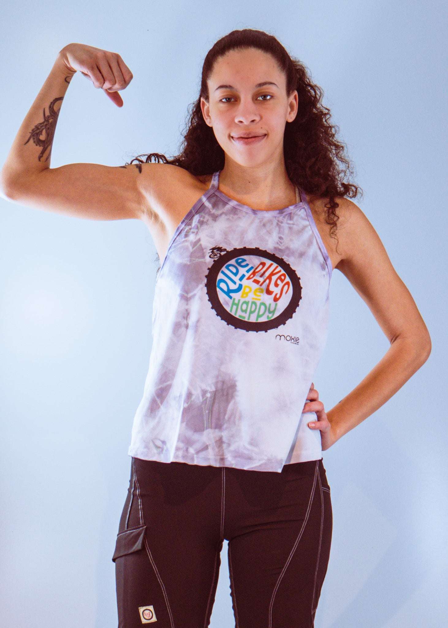 Woman wearing sleeveless tie dye bike top with "Ride Bikes Be Happy" graphic and black shorts flexing arm