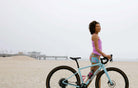 Woman wearing colorful gravel shorts standing with a bicycle on the beach.